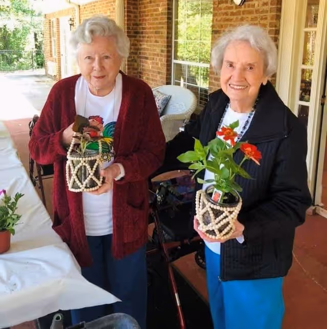 Two elderly women standing on a covered porch holding small potted plants decorated with beaded holders. They are smiling and dressed in casual clothing with cardigans. The porch has brick walls, windows, and outdoor furniture in the background.