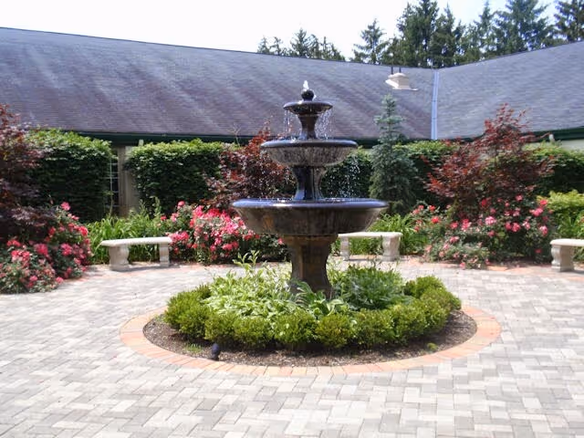 A courtyard with a three-tiered water fountain in the center surrounded by green shrubs and flowering plants. There are stone benches around the courtyard and a building with a sloped roof in the background.