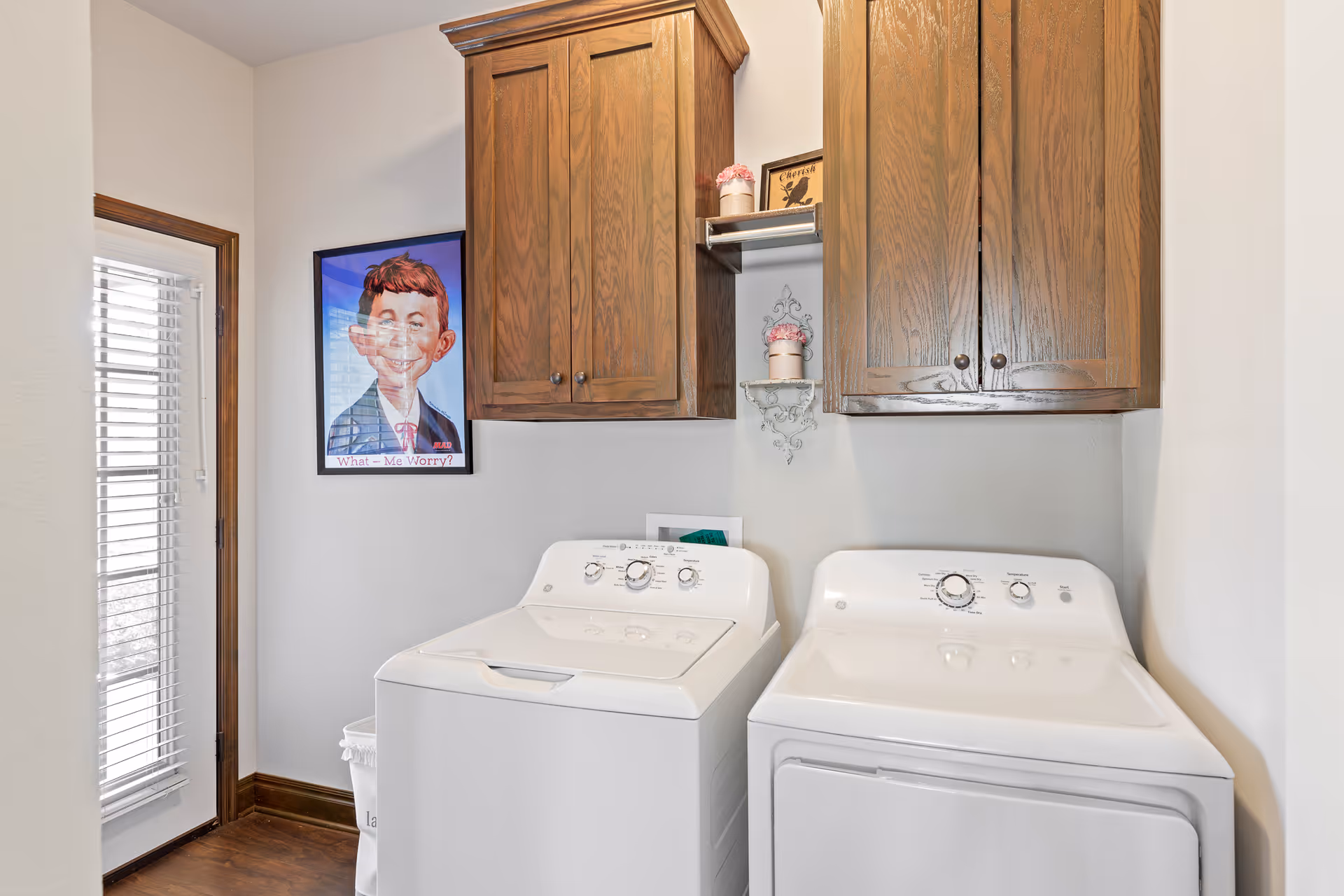Laundry room with a white washing machine and dryer side by side, wooden cabinets mounted on the wall above them, a framed cartoon poster on the wall, and a door with window blinds to the left.