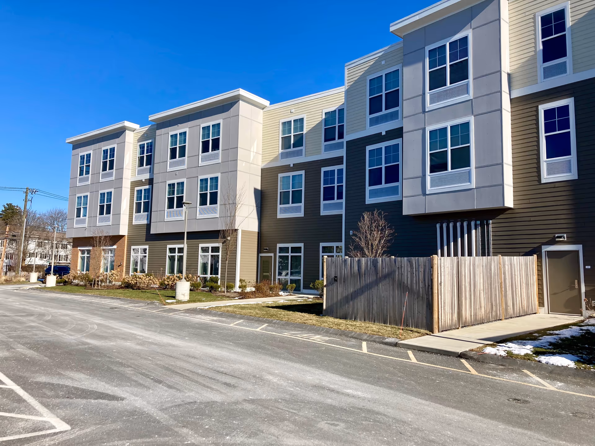 A modern three-story apartment building with multiple windows, a parking lot, and a wooden fenced area under a clear blue sky.