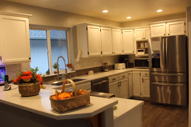 A modern kitchen with white cabinets, stainless steel refrigerator, dishwasher, and microwave. The kitchen features a large countertop with a basket of autumn leaves and a flower pot with red flowers. There is a window above the sink with a flexible faucet and some small plants on the windowsill. The floor is wooden and the kitchen is well-lit with recessed ceiling lights.
