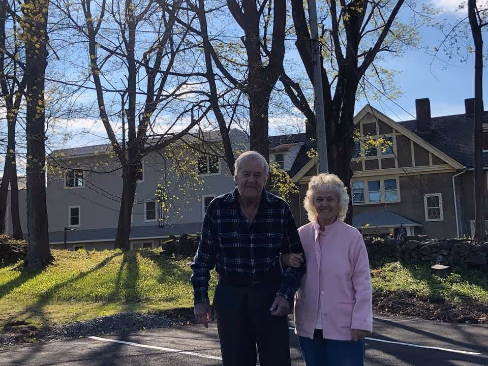 An elderly couple standing outside on a sunny day with trees and a large house in the background. The man is wearing a dark plaid shirt and dark pants, and the woman is wearing a light pink jacket and blue pants. They are smiling and holding arms.