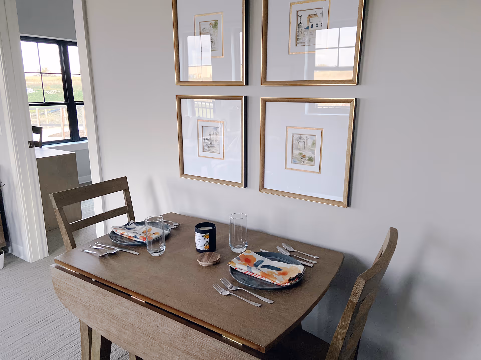 A small wooden dining table set for two with plates, colorful napkins, glasses, and cutlery. The table is positioned against a light gray wall with four framed pictures arranged in a square. A doorway to the left shows a glimpse of another room with large windows.