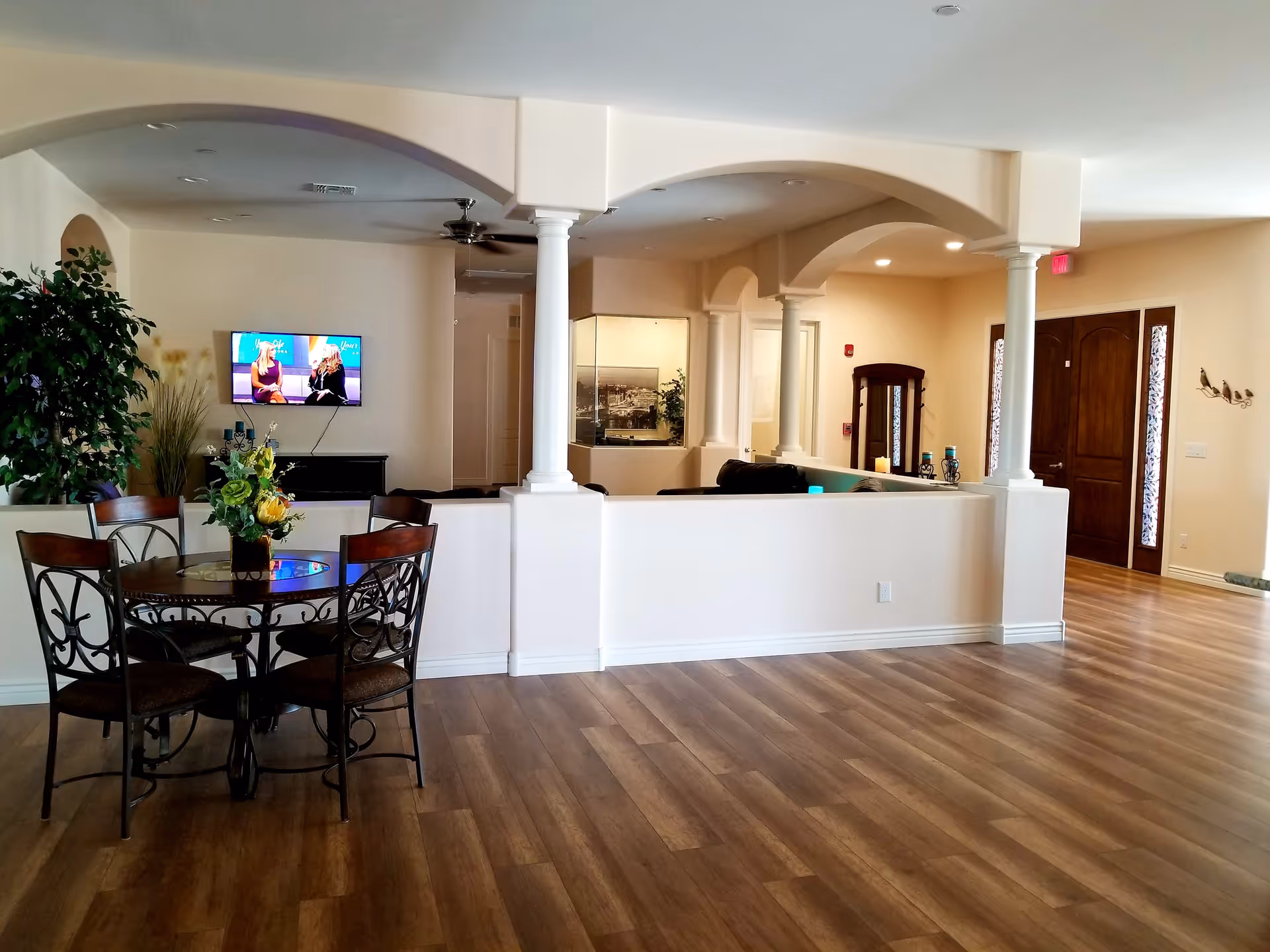 Interior view of a senior living facility common area with wooden flooring, a round glass-top table with four chairs and a floral centerpiece, a large potted plant, a wall-mounted TV showing two women talking, white half walls with columns and arches, and a wooden front door with sidelights.