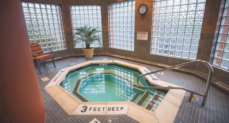 Octagonal indoor hot tub/spa in a tiled room with glass-block windows, a potted plant and chairs.