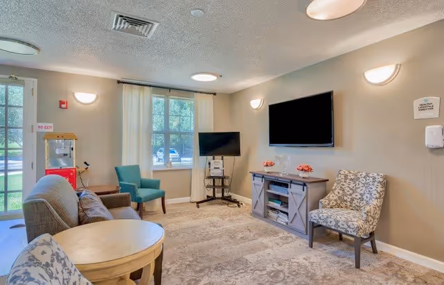 A cozy living room area with a patterned armchair, a teal armchair, a gray sofa, a round wooden table, a TV mounted on the wall above a wooden cabinet, and a smaller TV on a stand near a window with white curtains. There is a popcorn machine near the door with a 'NO EXIT' sign above it. The room is well-lit with wall sconces and ceiling lights.