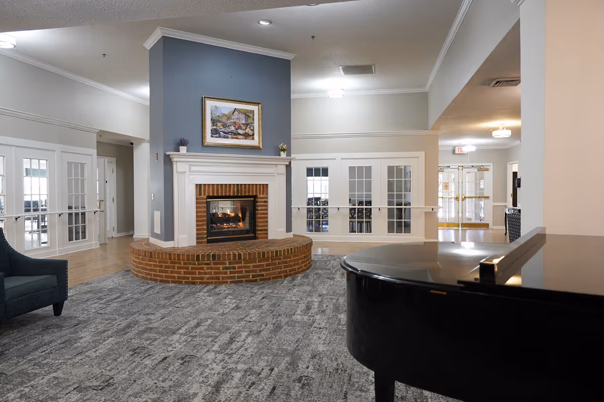 Common room with a brick fireplace and mantel topped by a painting, seating to the left and a grand piano in the foreground.