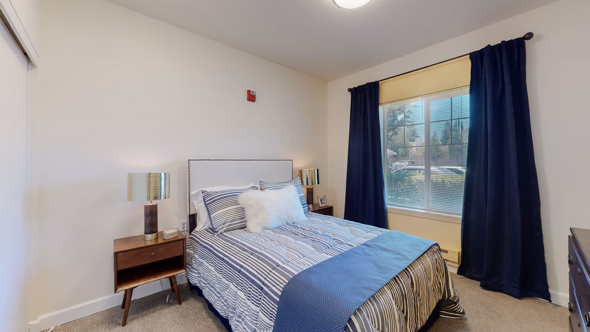 A bedroom with a bed dressed in blue and white striped bedding and a fluffy white pillow. There are two wooden nightstands on either side of the bed, each with a lamp. A large window with dark blue curtains and a beige blind lets in natural light. The walls are white and the floor is carpeted.