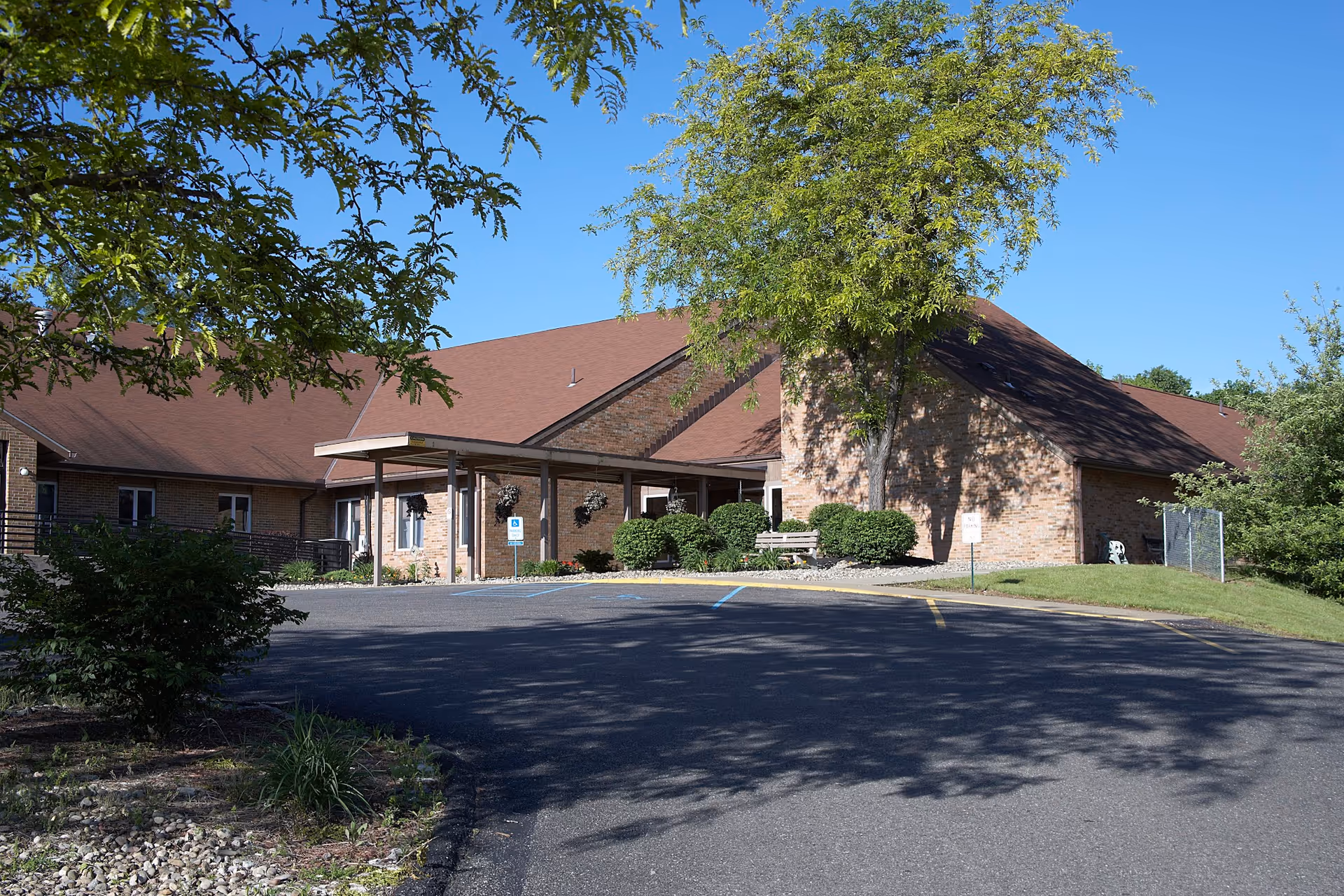 Exterior view of a single-story brick building with a brown roof, surrounded by trees and bushes, with a paved driveway and parking area in front under a clear blue sky.