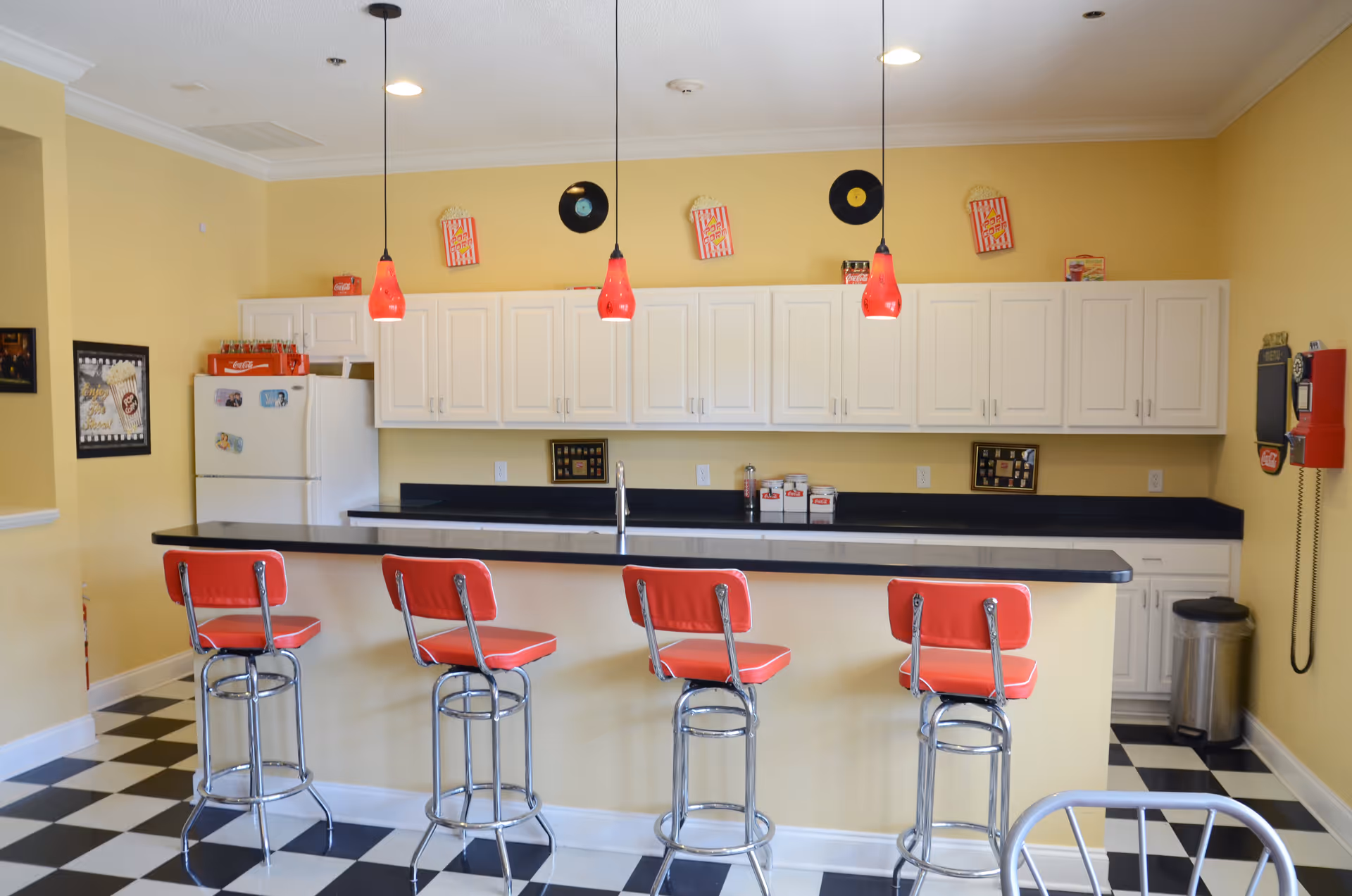 Retro-style kitchen/bar area with a black-and-white checkered floor, four red bar stools at a counter, white cabinets and a refrigerator under pendant lights.