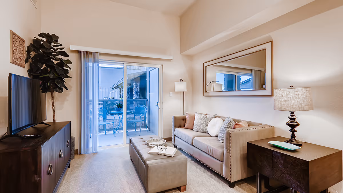 A cozy living room with a beige sofa adorned with multiple decorative pillows, a large rectangular mirror on the wall above the sofa, a dark wooden side table with a lamp, a matching ottoman with a book and glasses on it, a dark wooden TV stand with a flat-screen TV, a tall potted plant, and sliding glass doors leading to an outdoor patio with a small table and two chairs.