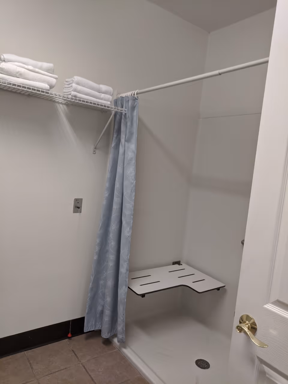 A small shower area with a light blue patterned shower curtain, a white fold-down shower seat, and a wire shelf above holding neatly folded white towels. The floor is tiled and the walls are white.