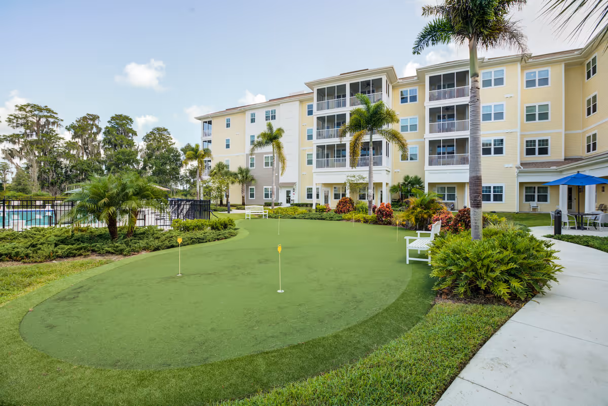Outdoor view of Arbor Terrace Citrus Park showing a putting green with small flags, surrounded by palm trees, bushes, and benches. In the background, there is a multi-story yellow and white building with balconies and windows under a partly cloudy sky.