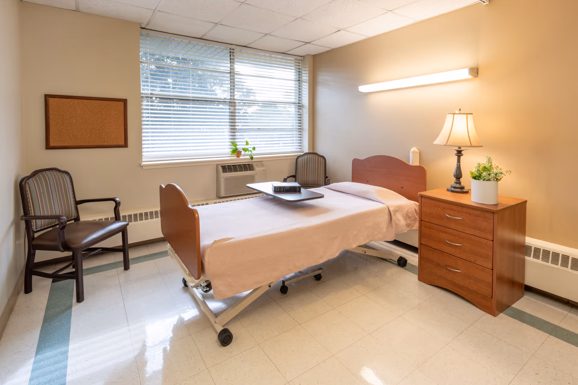 A tidy nursing facility bedroom with a single hospital bed, bedside table and two chairs by a window.