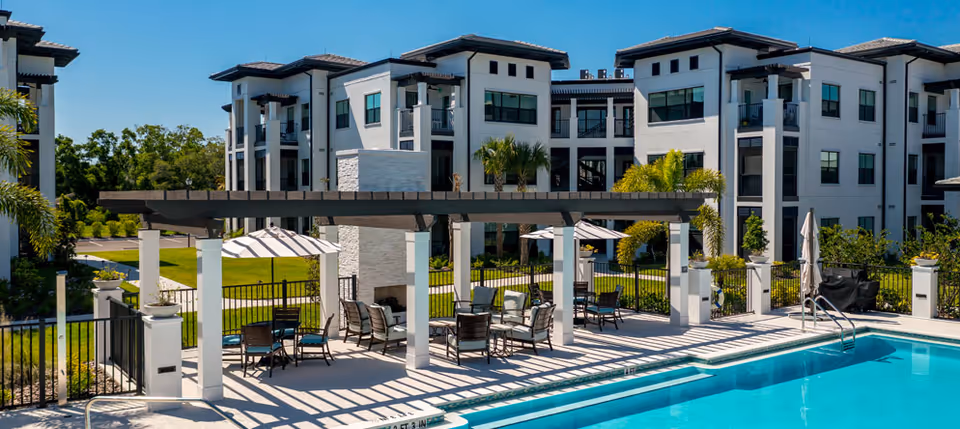 Outdoor pool area at Amira Choice Sarasota with a pergola providing shade over seating areas with tables and chairs, surrounded by a fence. In the background, there are modern three-story residential buildings with balconies and large windows, along with palm trees and greenery.