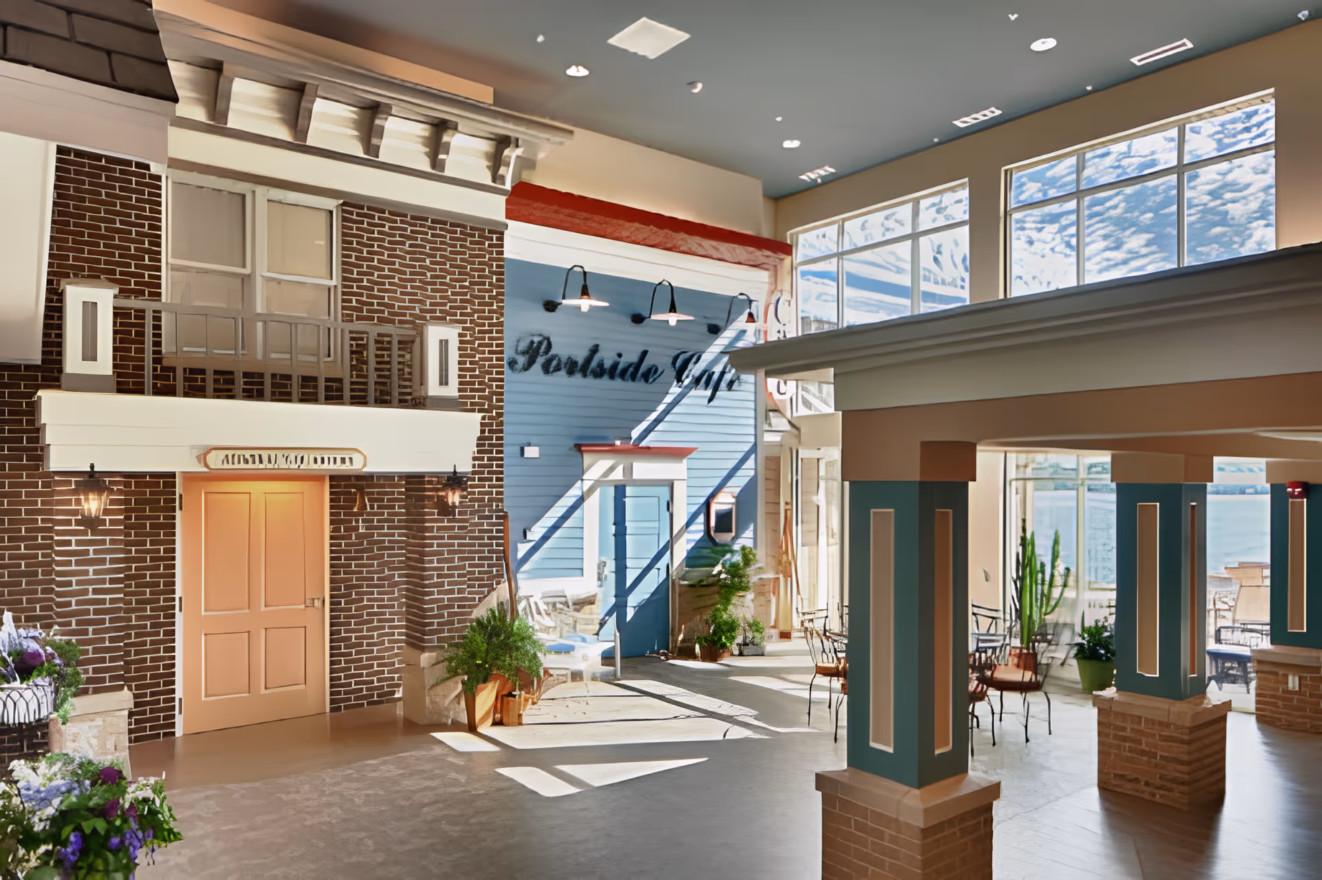 Bright indoor common area with large windows showing a view of a lake. The space features a brick wall with a peach-colored door, a blue wall with the words 'Poolside Cafe,' several potted plants, and metal chairs and tables arranged near the windows.