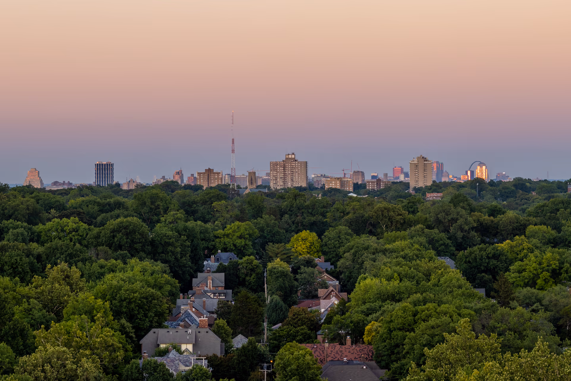 View of a city skyline at sunset with a dense forest and residential houses in the foreground under a gradient sky transitioning from light peach to blue.