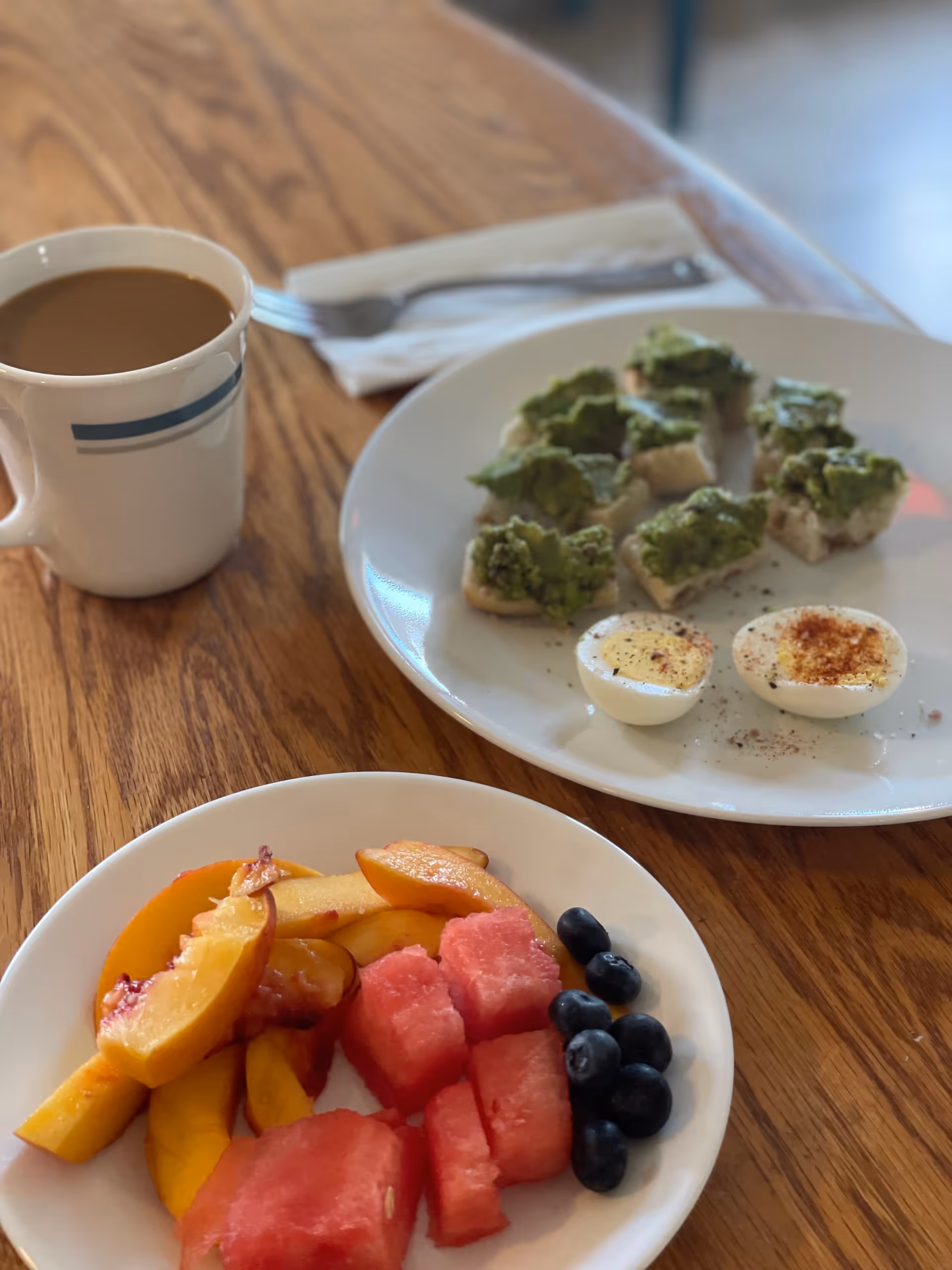 A wooden table with a white plate containing deviled eggs and small pieces of bread topped with green spread, a smaller white plate with sliced peaches, watermelon cubes, and blueberries, and a white mug filled with coffee. A fork and napkin are placed behind the larger plate.