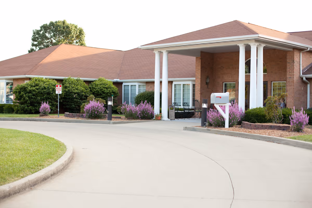 Driveway leading to the covered entrance of a brick senior living building with white columns, a mailbox, and landscaped flower beds.
