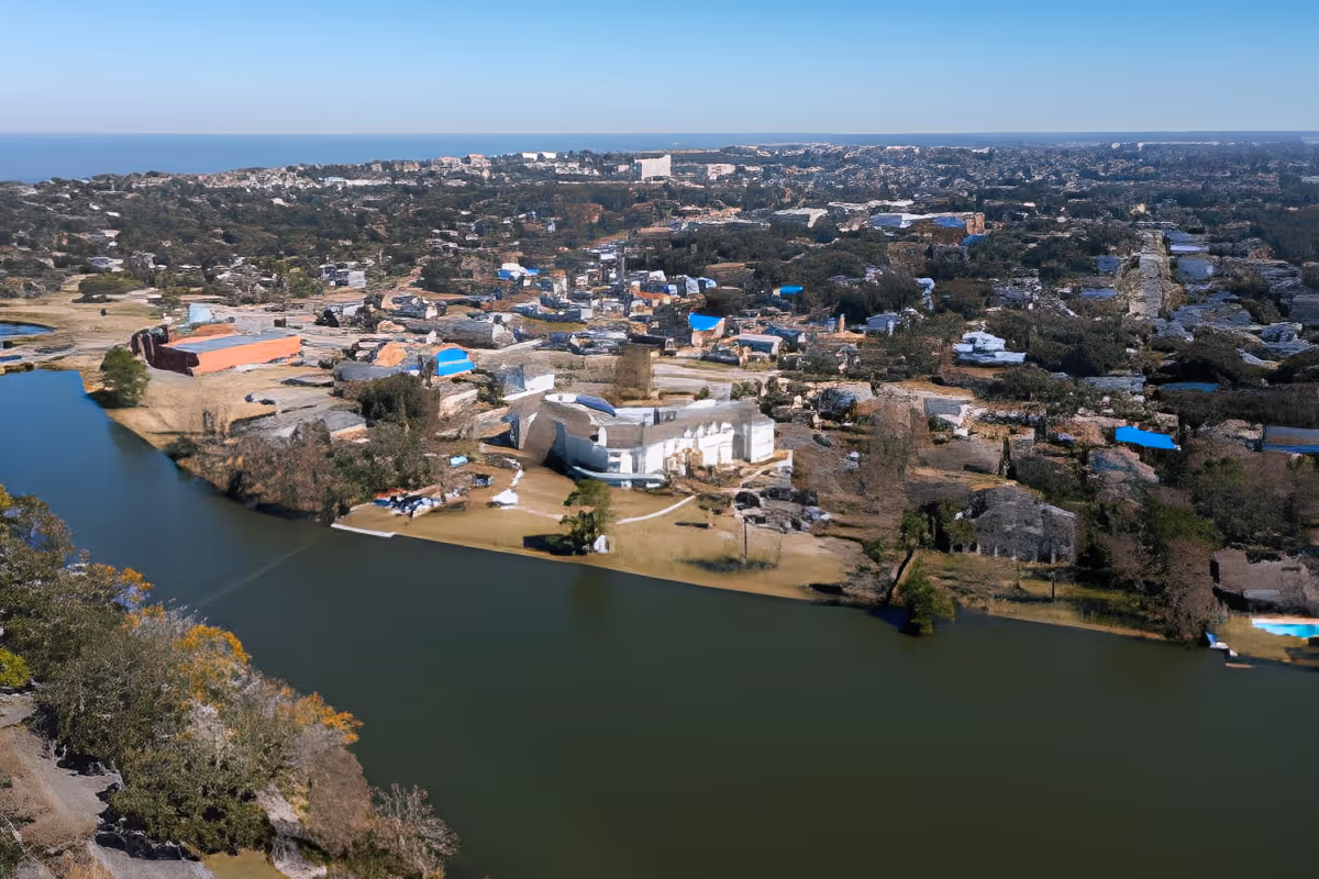 Aerial view of a residential area near a large body of water with trees and greenery surrounding the buildings under a clear blue sky.