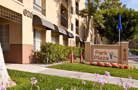 Exterior view of the Park Plaza senior living community building with beige walls, black awnings over windows, and the address number 620 displayed. A sign in front reads 'Park Plaza, A Kisco Senior Living Community' surrounded by green grass, flowers, and trees.