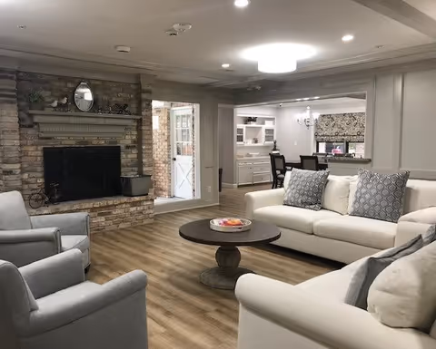A bright assisted living common room with sofas and armchairs arranged around a round coffee table and a brick fireplace, opening into a dining area.