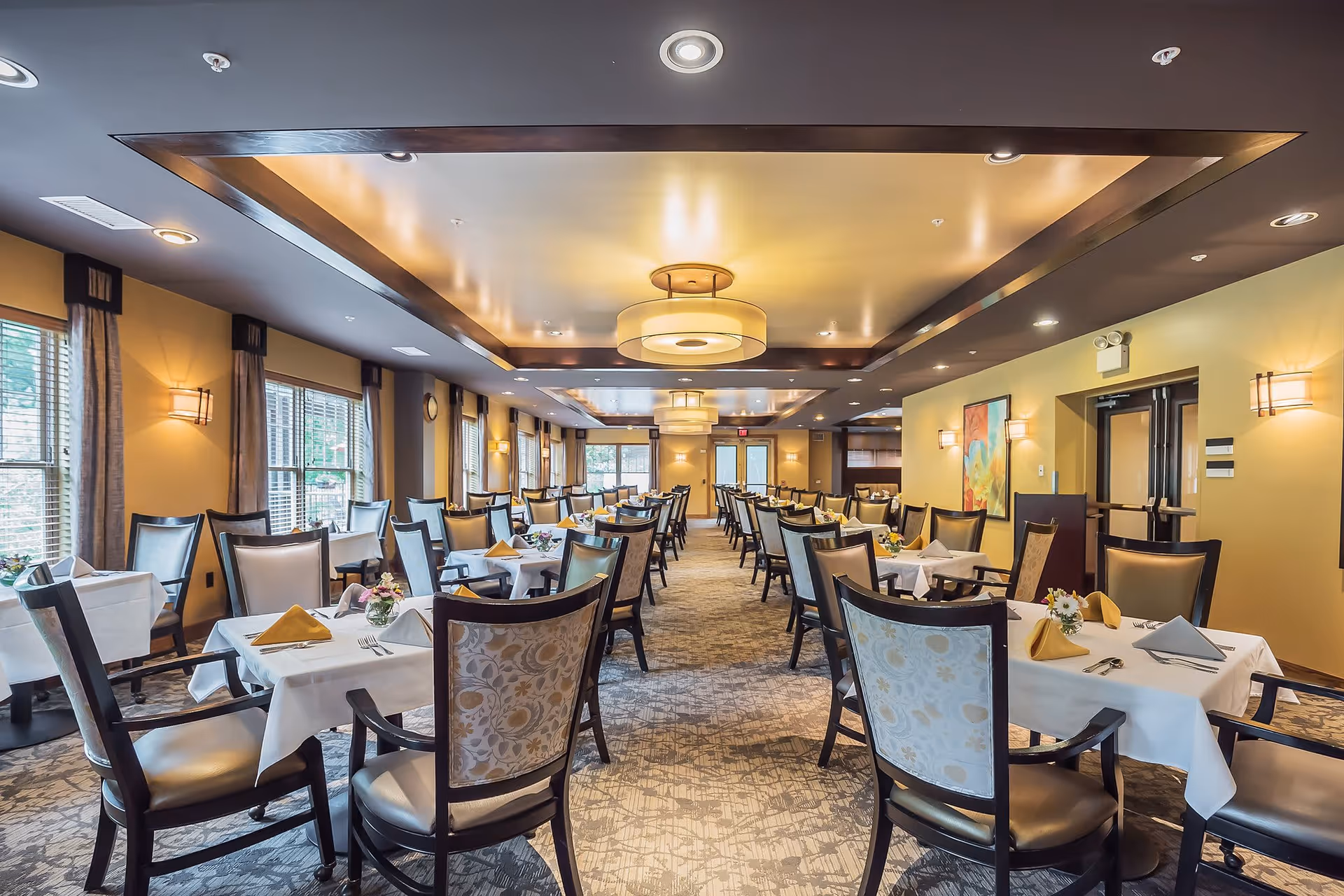 A spacious senior living dining room with multiple tables covered in white tablecloths, each set with napkins, cutlery, and small floral centerpieces. The room features large windows with curtains, warm yellow walls, carpeted flooring, and modern ceiling lights.