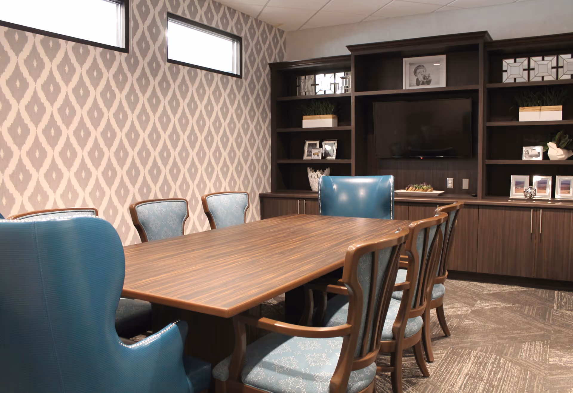 A conference or meeting room with a long wooden table surrounded by eight chairs, including two blue upholstered armchairs at the ends and six wooden chairs with blue patterned cushions. The room has patterned wallpaper, two small rectangular windows near the ceiling, and a large built-in dark wood shelving unit with a flat-screen TV and decorative items.