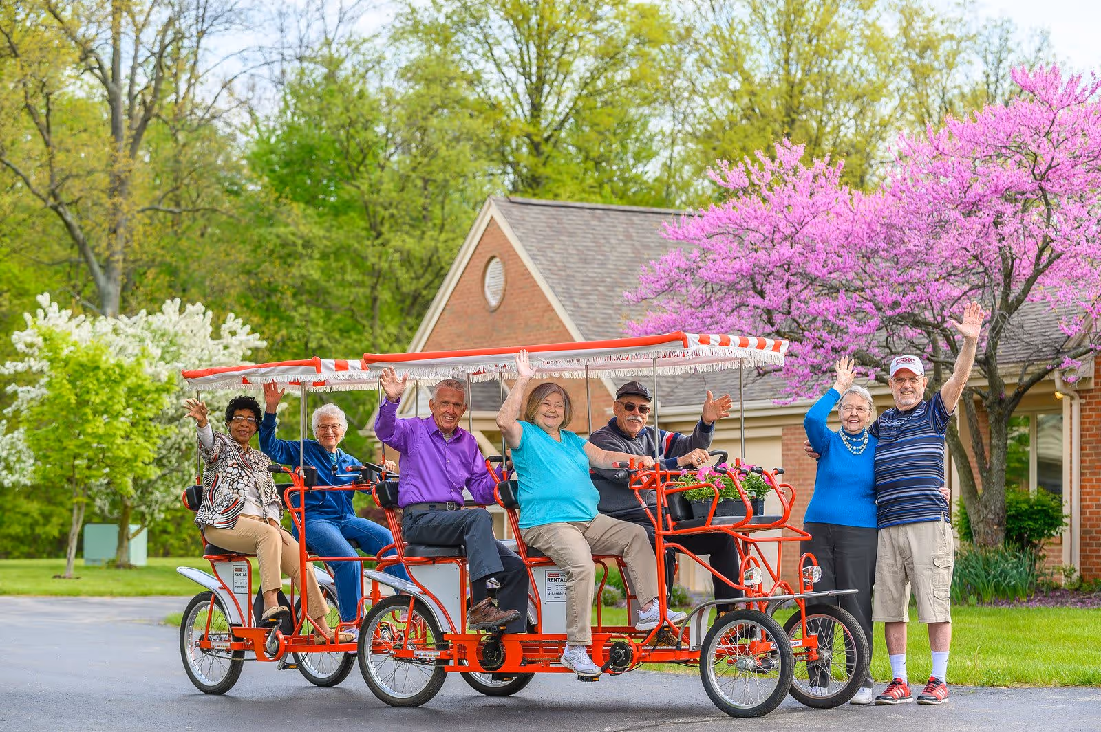 A group of six elderly people enjoying a ride on two red four-person pedal bikes with canopies, waving and smiling outdoors in front of a brick building with blooming trees and green foliage in the background.