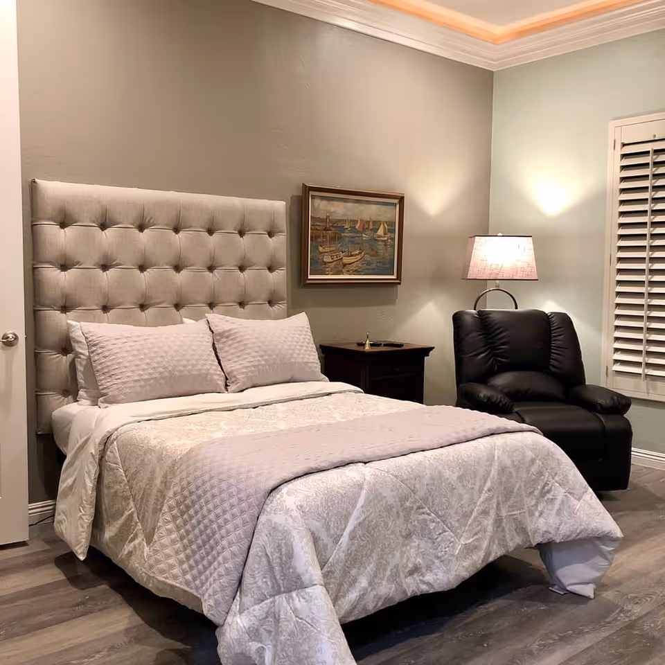A cozy bedroom with a tufted beige headboard, a neatly made bed with light-colored bedding and pillows, a dark wooden nightstand with a lamp, a black recliner chair, a framed painting of sailboats on the wall, and white plantation shutters on the window.
