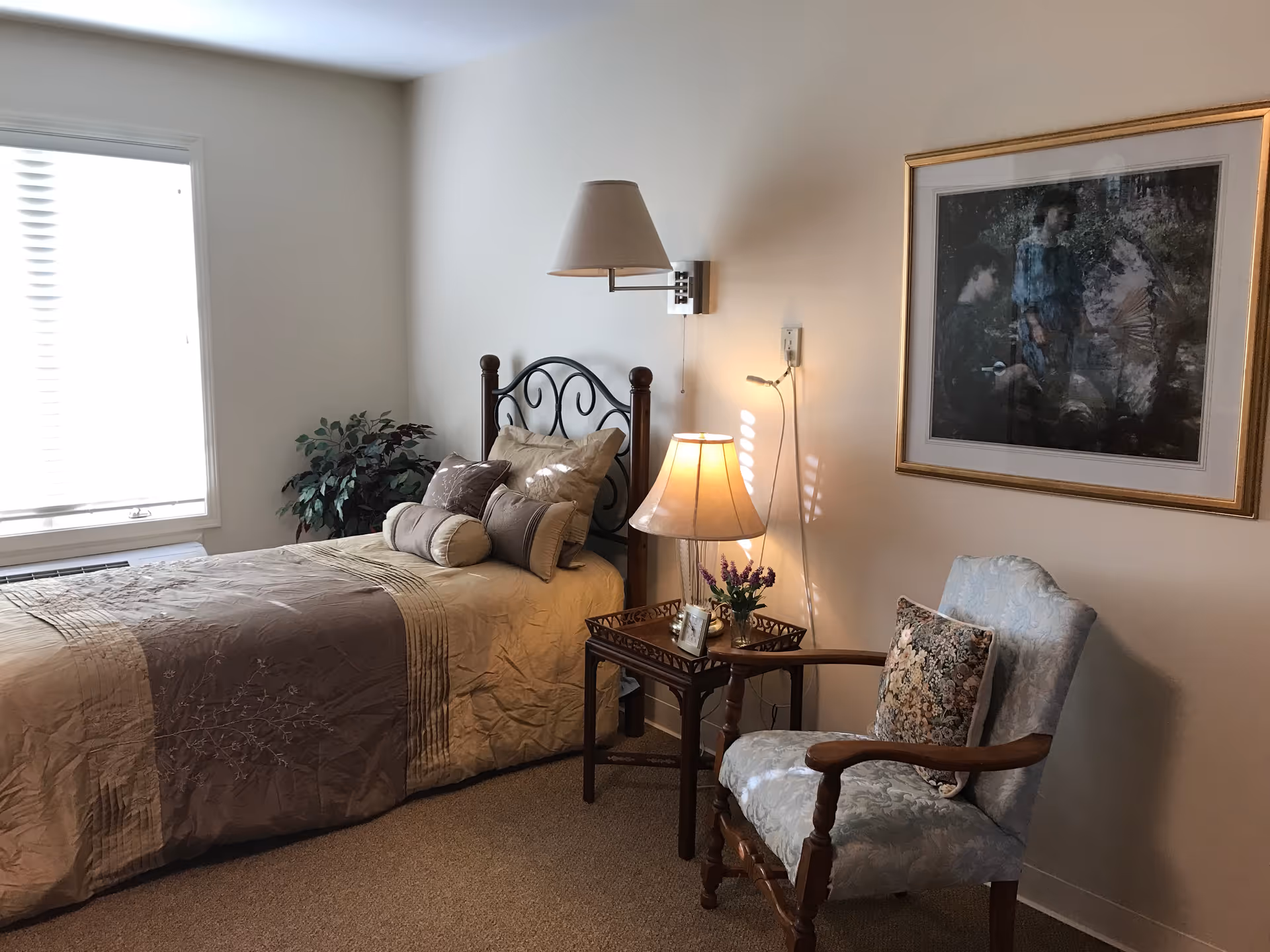 A cozy bedroom with a single bed covered in beige and brown bedding, multiple pillows, a wooden side table with a lamp and a small flower arrangement, a cushioned armchair with a floral pillow, a framed painting on the wall, and a window with blinds letting in natural light.