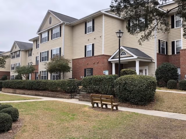 Front exterior of a three-story beige-and-brick apartment building with a lawn, walkway, bench, shrubs, and a covered entrance.