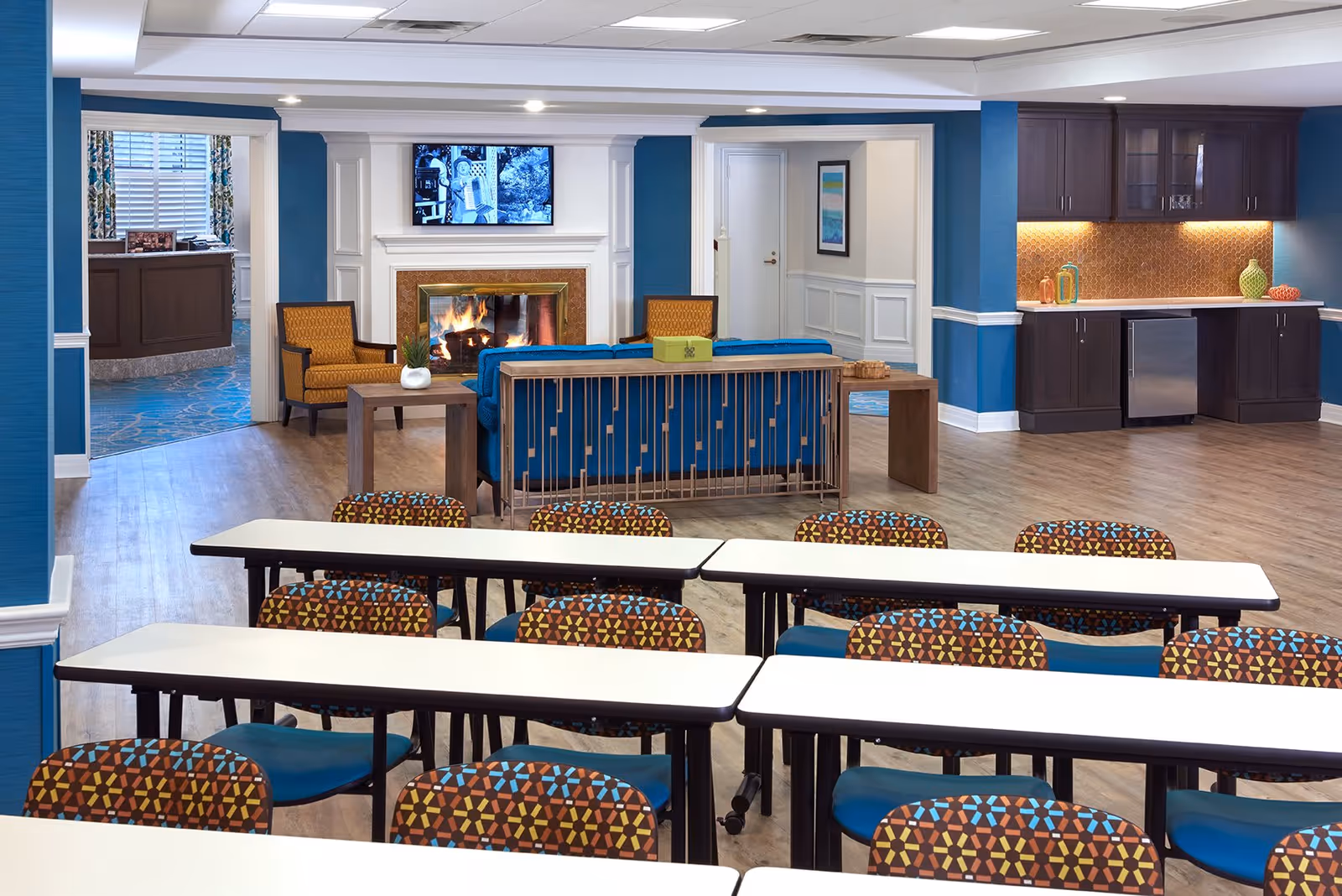 A spacious common area in an assisted living facility featuring rows of tables and colorful patterned chairs in the foreground. In the background, there is a cozy seating area with a blue sofa, two orange armchairs, a wooden coffee table, and a lit fireplace beneath a mounted TV. To the right, there is a kitchenette with dark wood cabinets, a small refrigerator, and decorative items on the countertop. The walls are painted blue with white trim, and the floor is light wood.