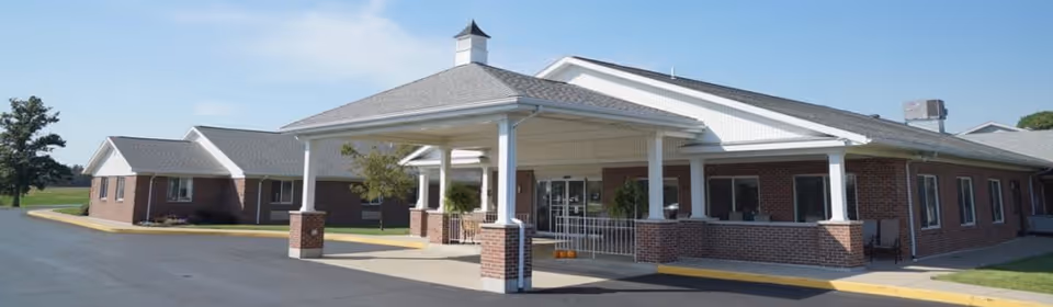 Exterior view of a single-story brick building with a covered entrance supported by white columns. The building has a gray shingled roof and a small cupola on top. There is a paved driveway and some greenery around the building under a clear blue sky.