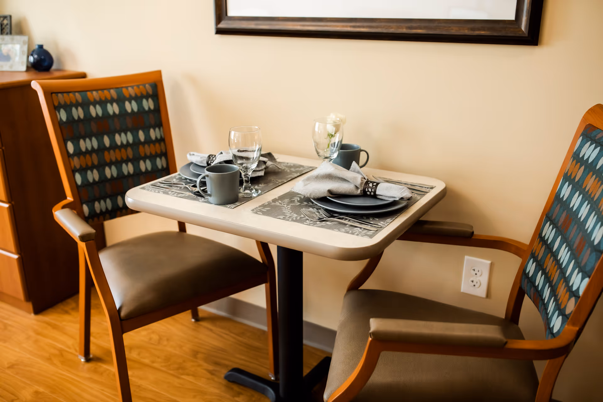 A small dining table set for two with two chairs featuring patterned backs and cushioned seats. The table has placemats, plates, napkins with rings, mugs, and glasses. The setting is indoors with a wooden floor and a framed picture on the wall.