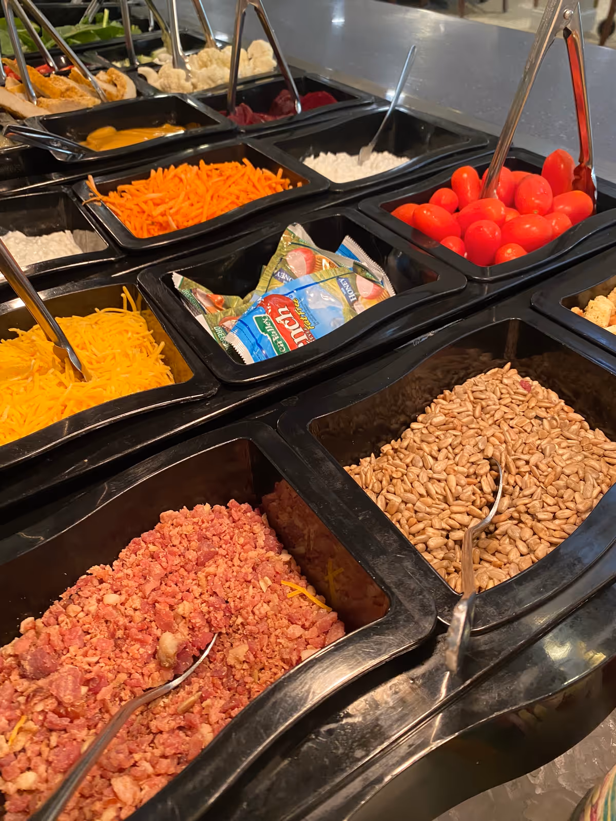 A salad bar with black bins holding shredded cheese, bacon bits, sunflower seeds, cherry tomatoes, shredded carrots and dressing packets.