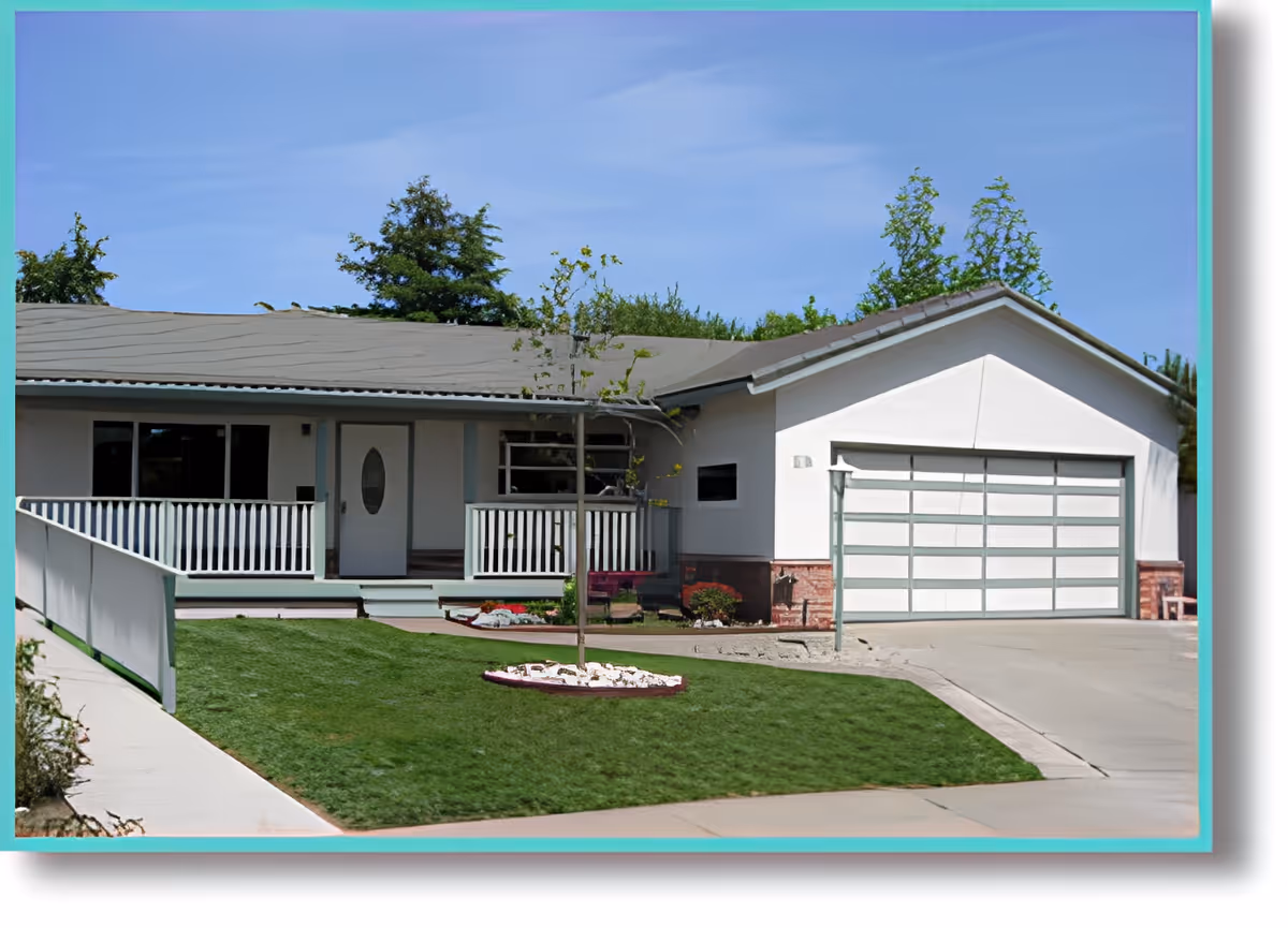 Front exterior view of a single-story house with a white facade, a gray roof, a two-car garage, a small porch with white railings, a young tree planted in the front yard, and a well-maintained lawn under a clear blue sky.