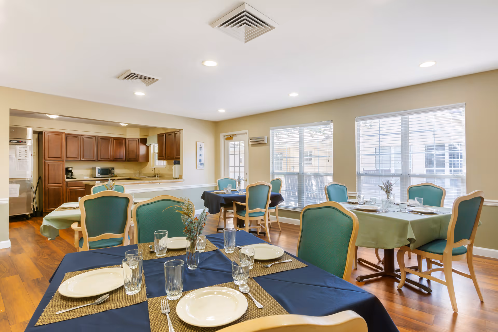 A bright dining room in a senior living facility with several tables covered in blue and green tablecloths, set with plates, glasses, and silverware. The room has large windows letting in natural light, wooden flooring, and a view into a kitchen area with wooden cabinets.