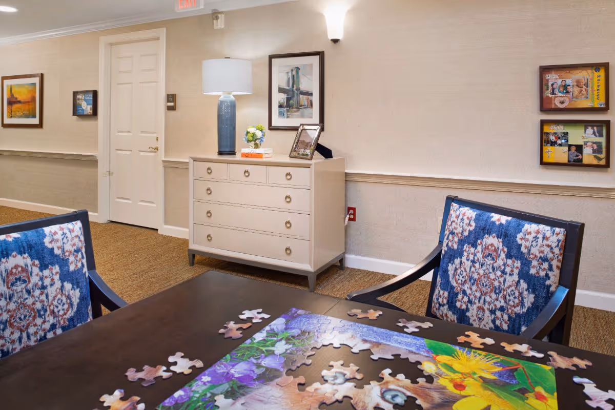 A cozy interior room with a dark wooden table featuring a partially completed colorful jigsaw puzzle. Two chairs with blue and white floral upholstery are positioned at the table. Against the wall, there is a light-colored dresser with a blue lamp, a small flower arrangement, and a framed photo. The walls are decorated with framed pictures and a bulletin board with photos. The floor is carpeted, and a closed white door is visible in the background.