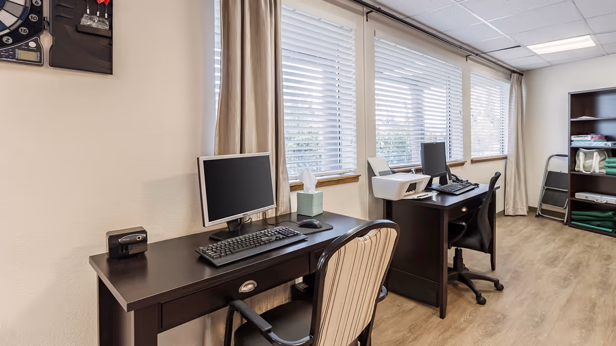 A well-lit office area with two dark wood desks placed side by side along a wall with large windows covered by white blinds. Each desk has a computer monitor, keyboard, and mouse, with a tissue box on one desk and a printer on the other. There are two office chairs, one with striped upholstery and the other a black swivel chair. In the background, there is a dark wood shelving unit with folded green blankets and other items, and a small step ladder leaning against the wall. The floor is light wood, and the ceiling has recessed lighting.