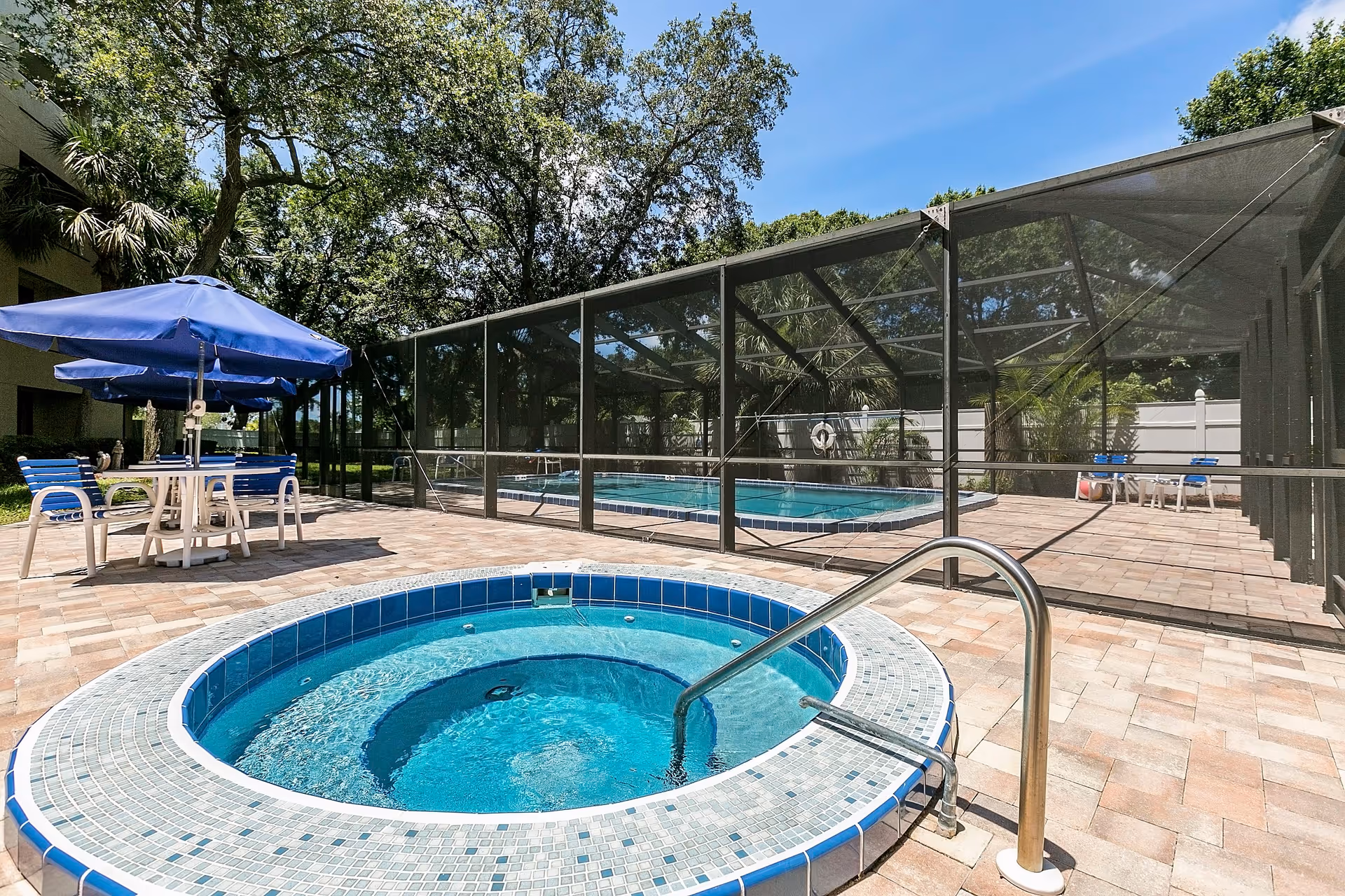 Outdoor area at Belleair Towers featuring a round hot tub with blue mosaic tiles in the foreground, a screened-in rectangular swimming pool in the background, and a patio with blue and white chairs and tables under blue umbrellas. Trees and a clear blue sky are visible.