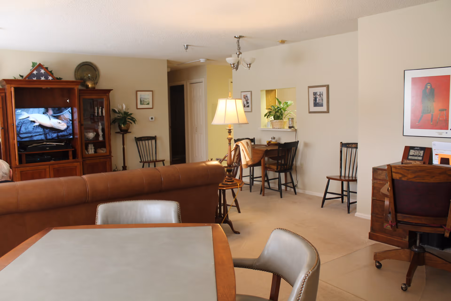 A cozy living room area with a brown leather sofa facing a wooden entertainment center with a TV showing a fish. There are several chairs, a floor lamp, framed pictures on the walls, a small desk with a chair, and a dining table with chairs in the foreground. The room has beige walls and carpeted floors.