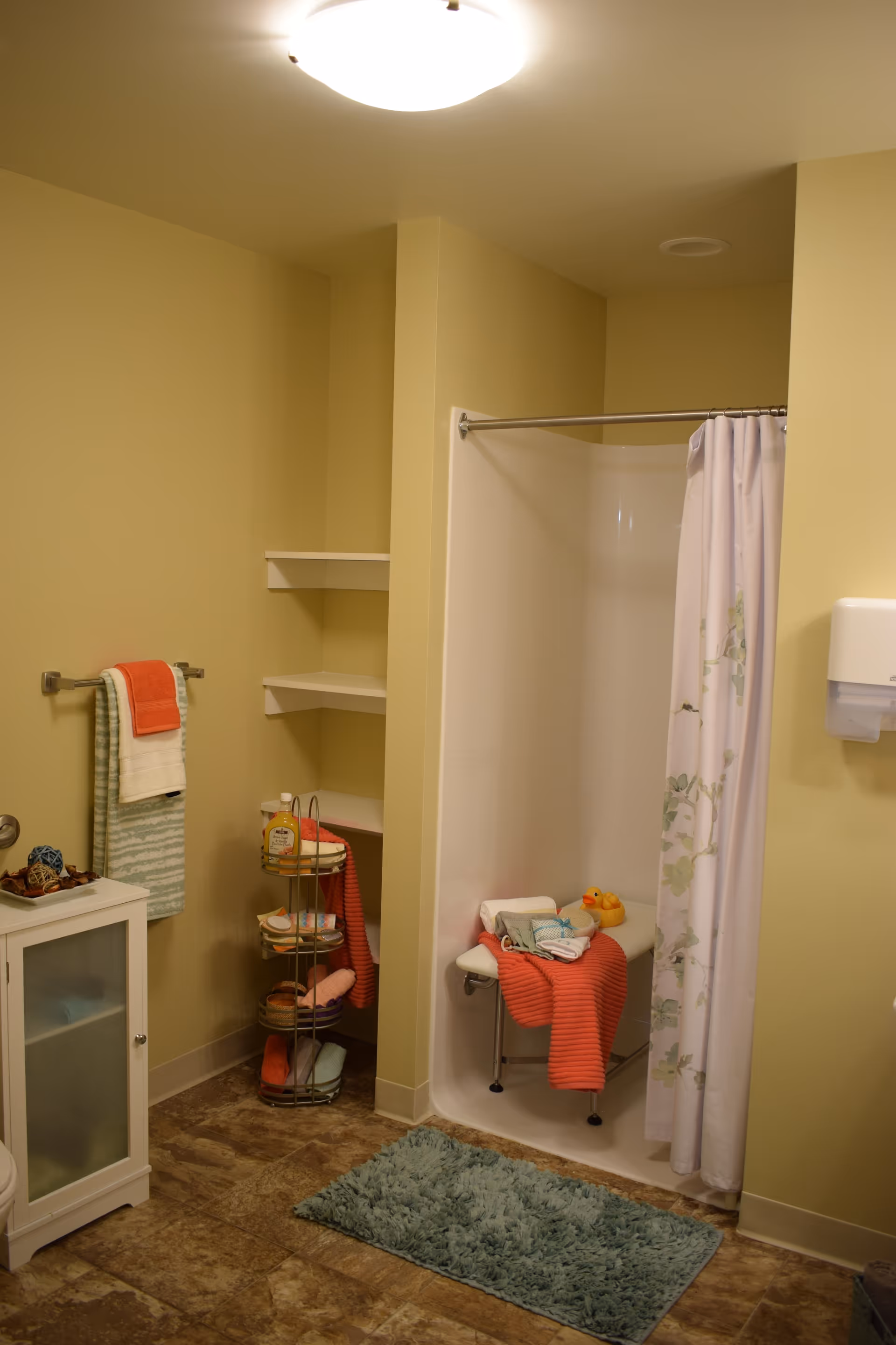 A bathroom with a shower area that has a white curtain with a floral pattern. Inside the shower is a white bench with folded towels and two rubber ducks on it. The bathroom has beige walls, a towel rack with two towels, a small white cabinet, a corner shelf with various toiletries, and a blue bath mat on the floor.