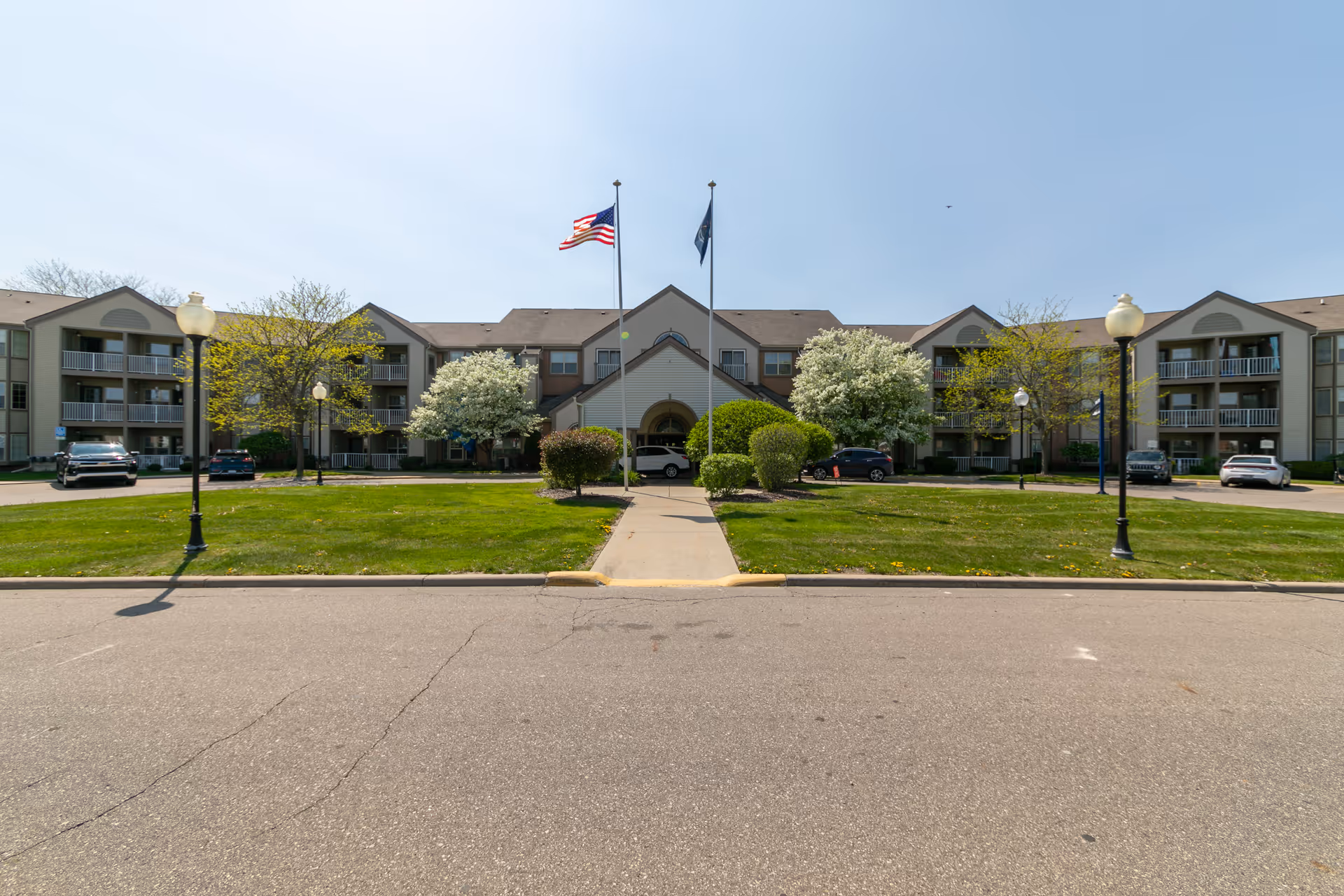 Front exterior of a multi-story senior living facility with flags, a central entrance, landscaped lawn, walkway, and parked cars.