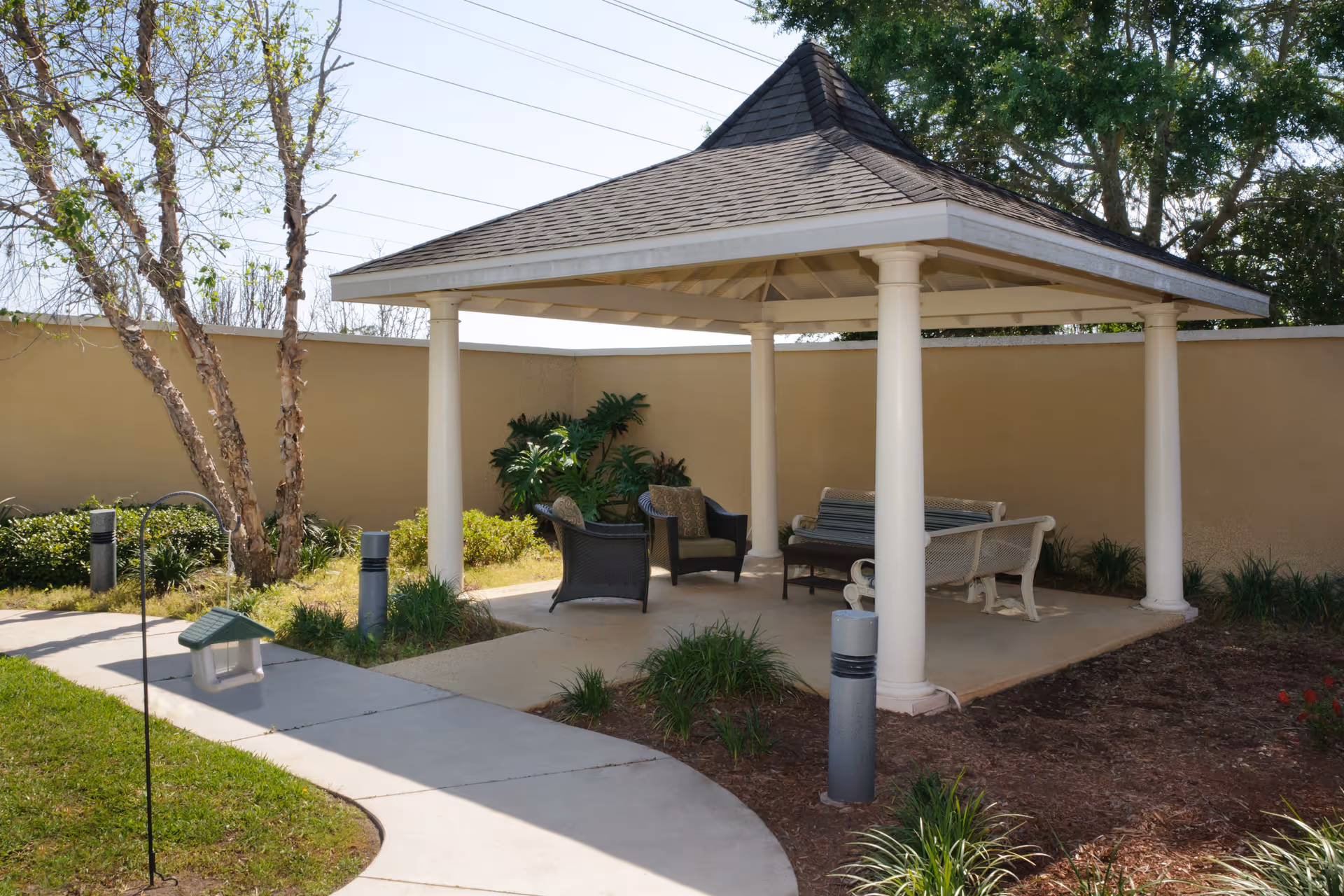 Outdoor gazebo with a shingled roof supported by white columns, containing wicker chairs and benches, surrounded by a concrete pathway, grass, plants, and a beige privacy wall.