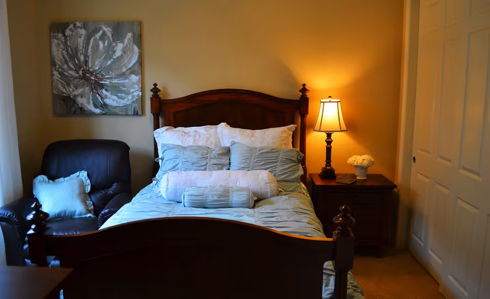A cozy bedroom with a wooden bed frame, neatly made bed with light blue and white pillows and bedding, a wooden nightstand with a lamp and a small flower pot, a dark leather armchair with a light blue pillow, and a large floral painting on the wall.