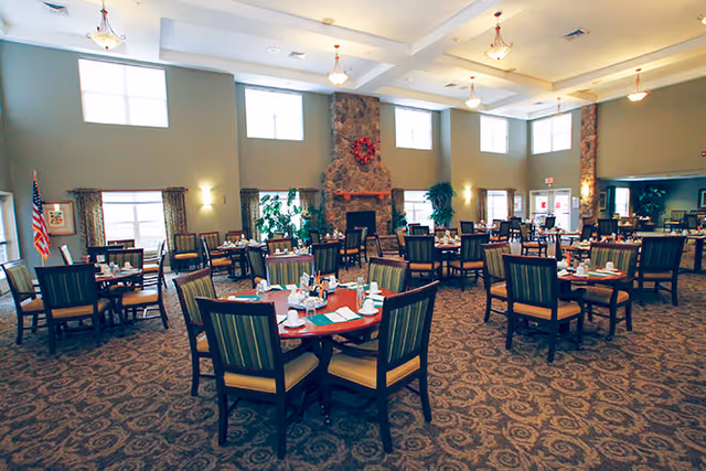 Large dining room with round tables set for meals, upholstered chairs, high ceilings, and a central stone fireplace.