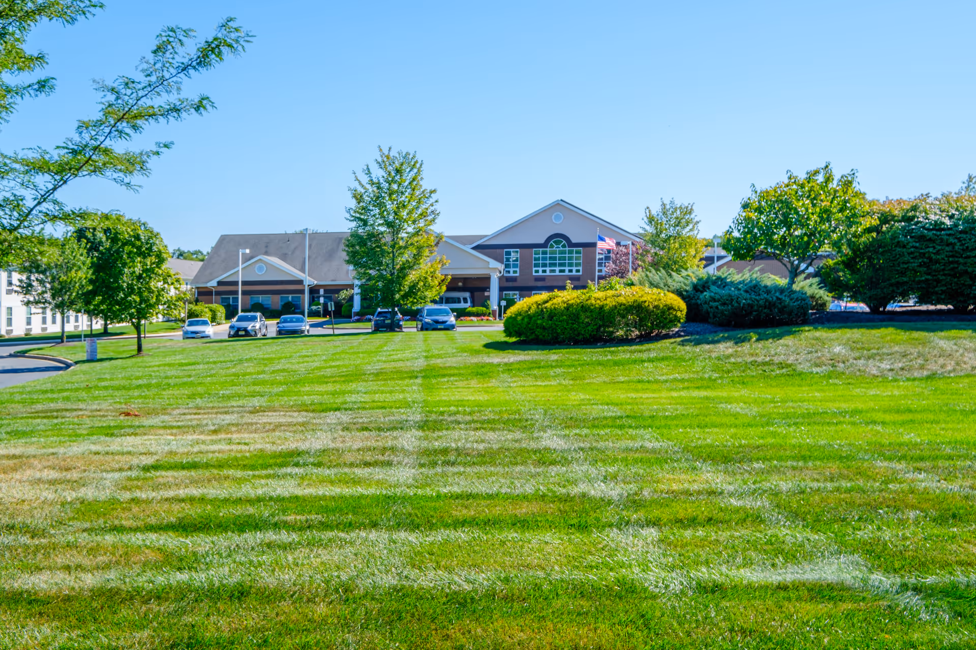 Wide view of a senior living facility building with a well-maintained green lawn in the foreground, several trees and bushes, and a few parked cars near the entrance under a clear blue sky.