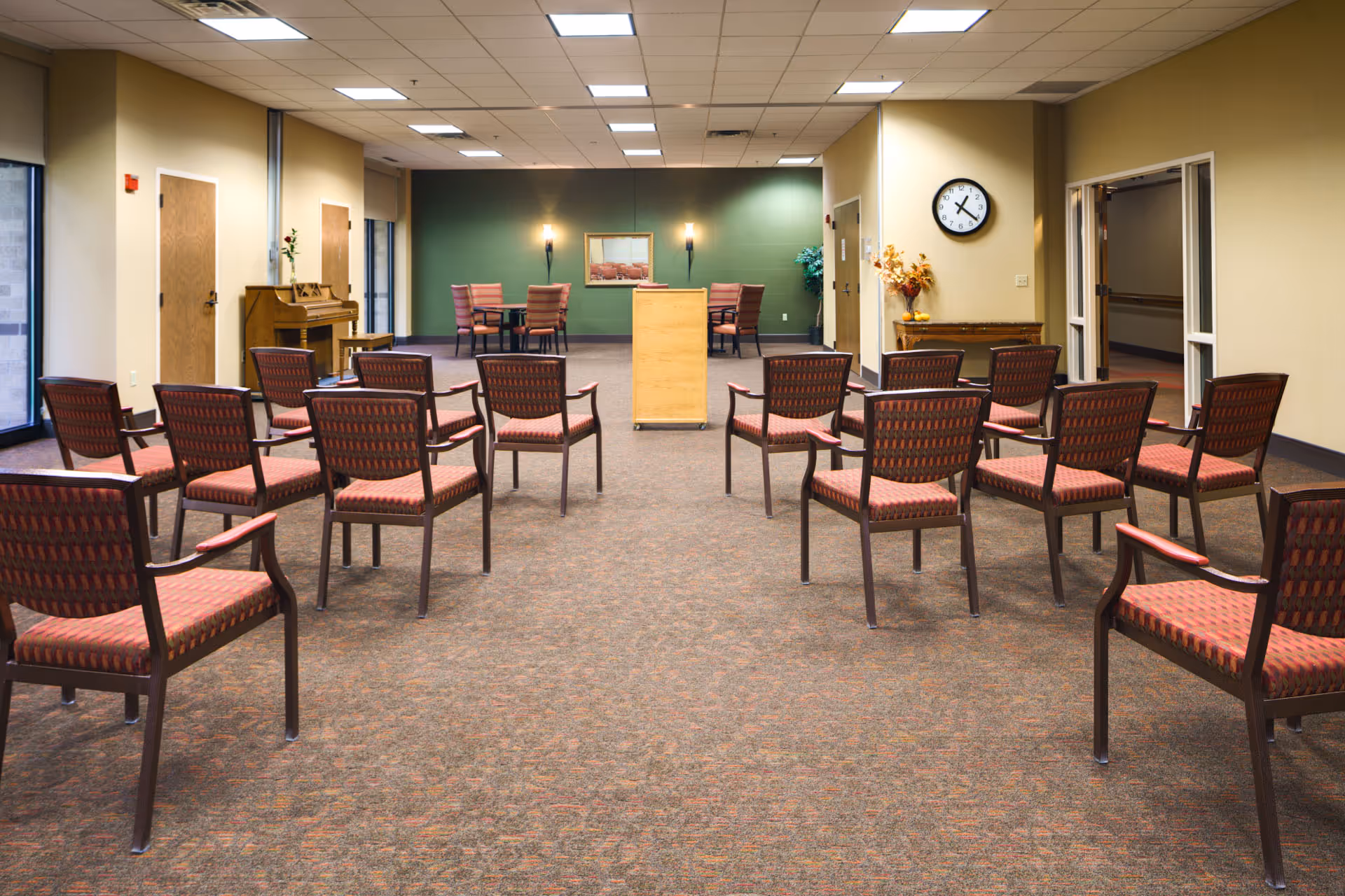 Empty activity/meeting room with rows of red-upholstered chairs facing a wooden podium and a piano.