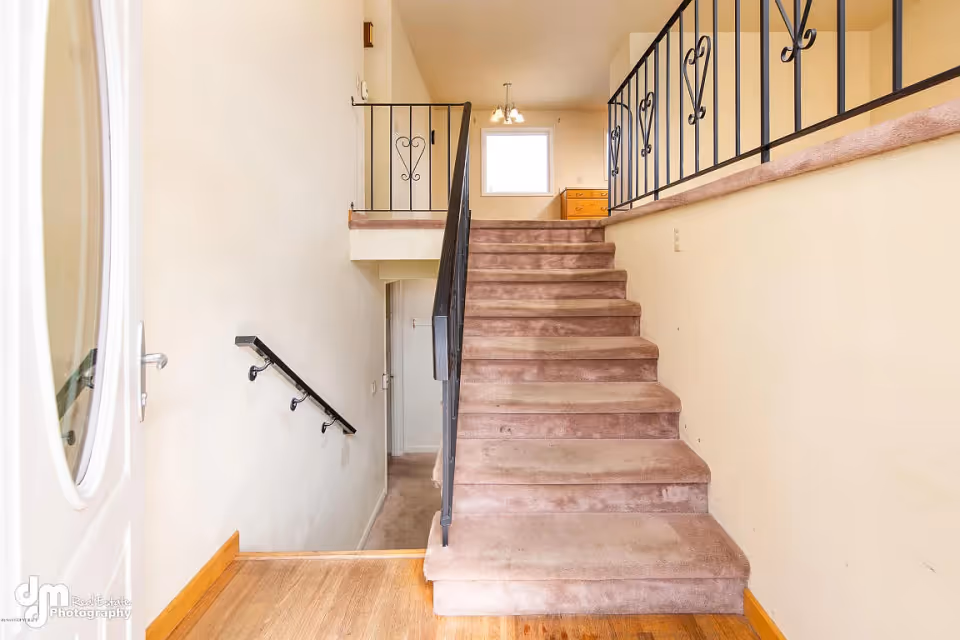 Interior view of a stairway with carpeted steps leading up to a landing with a window and a wooden dresser. The stair railing is black metal with decorative heart-shaped designs. The walls are painted light beige, and there is a wooden floor at the base of the stairs.